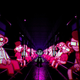 A pink glow shines off the skin of train car passengers who surround the game's main character in a dark train car while brandish creepy smiles.